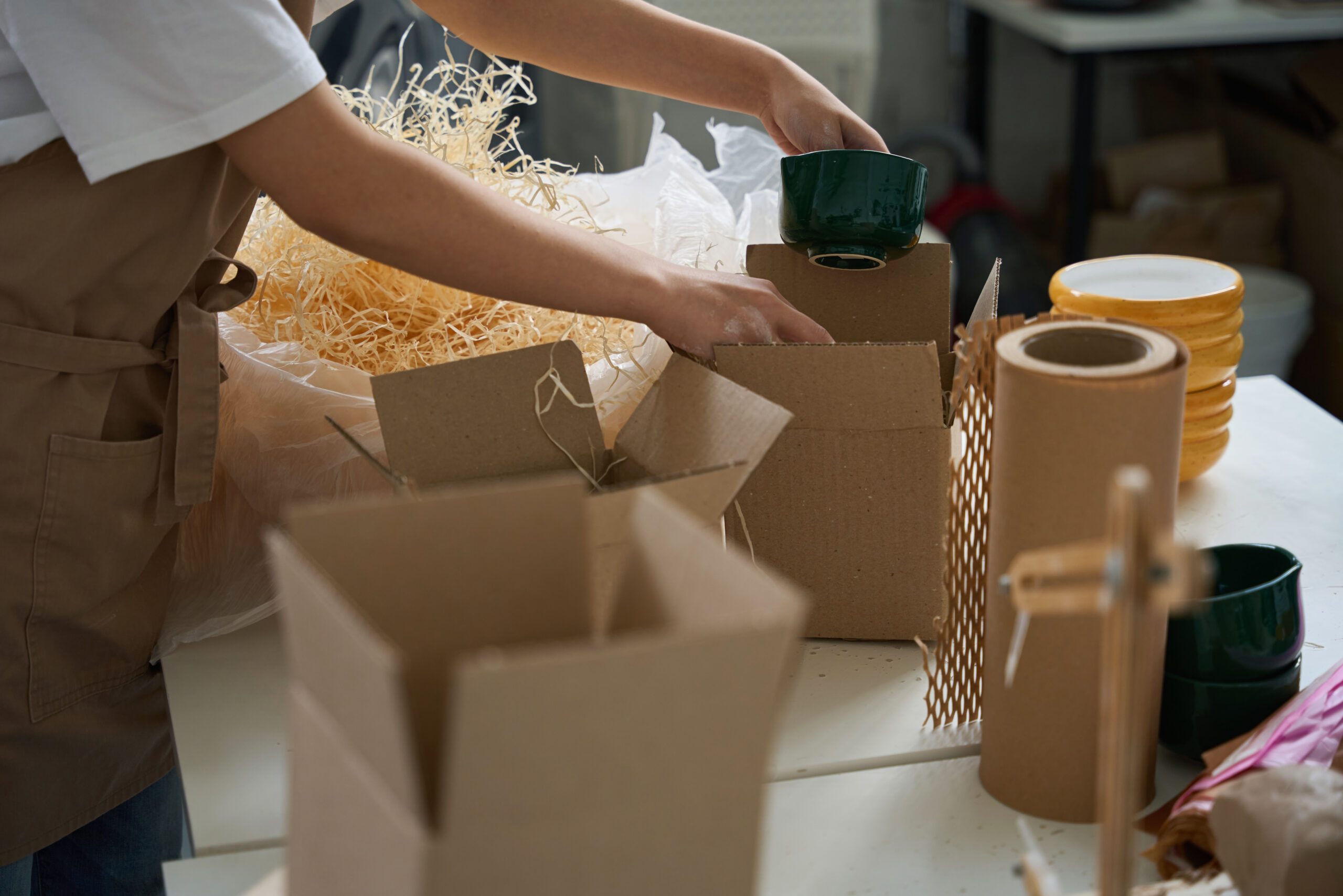 Young woman is packing a beautiful ceramic cup into a cardboard box, packing material and twine are nearby